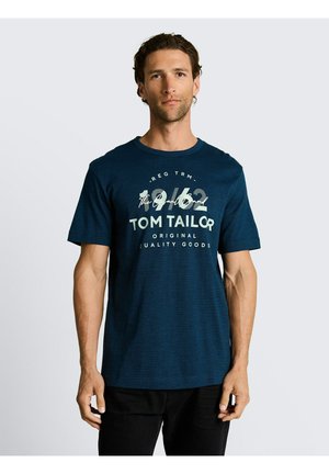 Mężczyzna ubrany w granatowy t-shirt Tom Tailor z białym napisem i cyframi, stojący na jednolitym jasnoszarym tle.