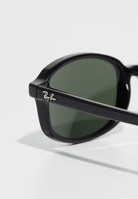 Gafas de sol de plástico negro con lentes verdes, con forma rectangular y un logo plateado de Ray-Ban en la patilla.