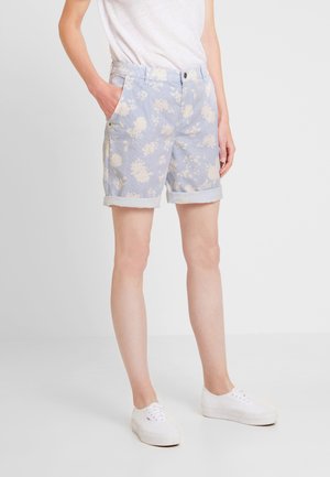 Persoon die lichtblauwe korte broek met opgerolde zoom en bloemenpatroon draagt, een wit T-shirt en witte sneakers, staand met één hand in de zak tegen een effen achtergrond.