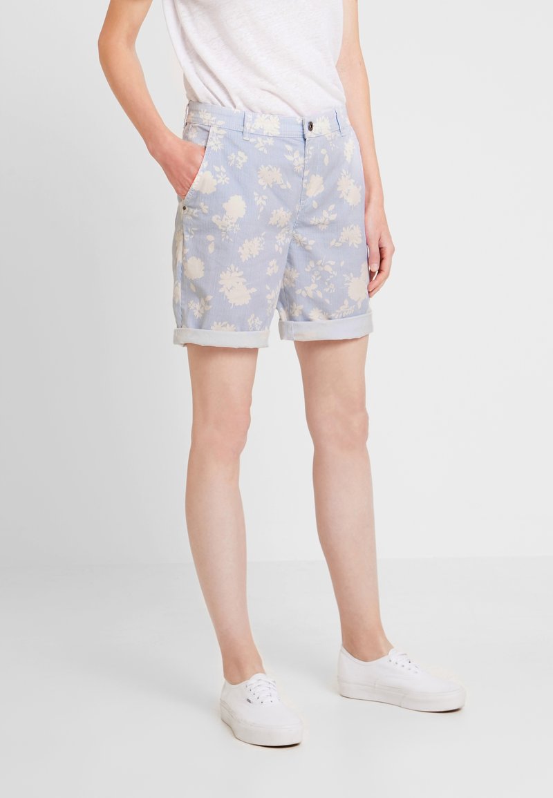 Personne portant un short bleu clair à fleurs avec ourlet roulé, t-shirt blanc, baskets blanches, debout avec une main dans la poche sur un fond uni.