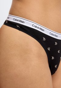 Μαύρο βαμβακερό στρινγκ με λευκή ελαστική μέση που φέρει το λογότυπο "Calvin Klein". Σημαντικά μοτίβα με το μονόγραμμα CK σε αντίθετα χρώματα.