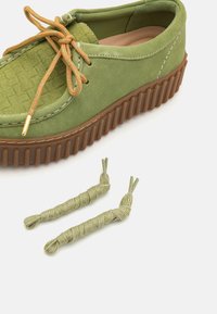 Zapato verde de ante con parte superior texturizada, cordones a juego y suela de goma marrón. Incluye dos cordones adicionales envueltos en un color similar.