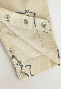 Beige kordflorsfabric med svarta djurkonturer och silverfärgade tryckknappar, som visar insidan och den vikta kanten av plagget.