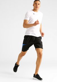 Wit sportshirt met korte mouwen, zwarte korte broek met blauwe accenten, zwarte hardloopschoenen; atleet te zien in het midden van de run.