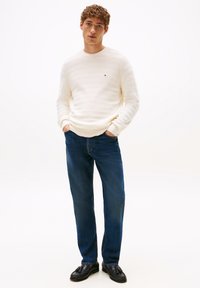 Gestreifter cremefarbener Pullover, blaue Jeans und schwarze Slipper. Der Pullover hat einen Rundhalsausschnitt und gerippte Bündchen. Das Model steht mit den Händen in den Taschen.