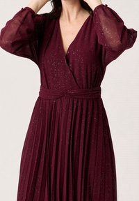 Burgunder Kleid mit transparenten, puffärmeln, einem tiefen V-Ausschnitt und einem plissierten Rock. Der Stoff weist ein dezentes, glitzerndes Muster auf.