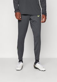 Grijze sportbroek met een slim fit, voorzien van een klein neon geel logo. Gepaarde met witte voetbalschoenen en zwarte sokken.