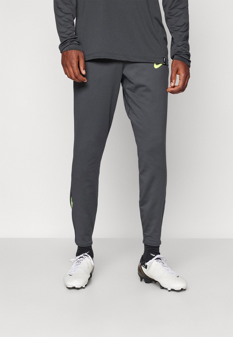 Grijze sportbroek met een slim fit, voorzien van een klein neon geel logo. Gepaarde met witte voetbalschoenen en zwarte sokken.