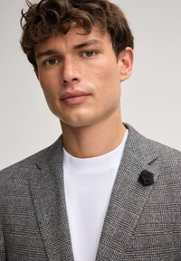 Grijze geruite blazer van textuurstof, met een zwarte bloemenpin op de revers. Draag het over een wit T-shirt met ronde hals.