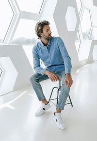 Svetlomodrá denimová košela, slim-fit svetlé džínsy a biele tenisky s čiernymi akcentmi. Model sedí na kovovom stoličkách v jasnom, modernom prostredí.