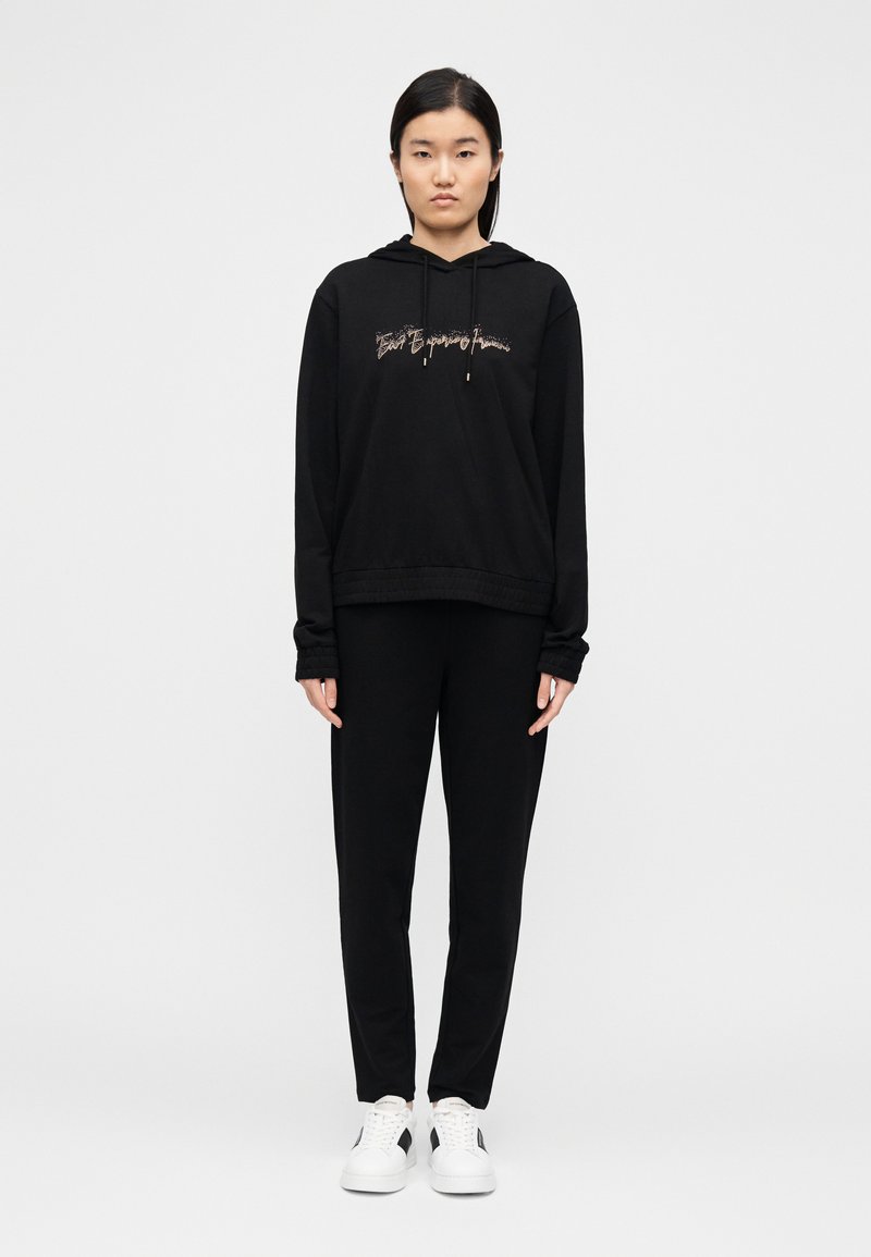 Μαύρο φούτερ με κουκούλα και κορδόνια, με γραφικό κείμενο, συνοδευόμενο από μαύρο παντελόνι και λευκά αθλητικά παπούτσια. Λείο ύφασμα, άνετη εφαρμογή.