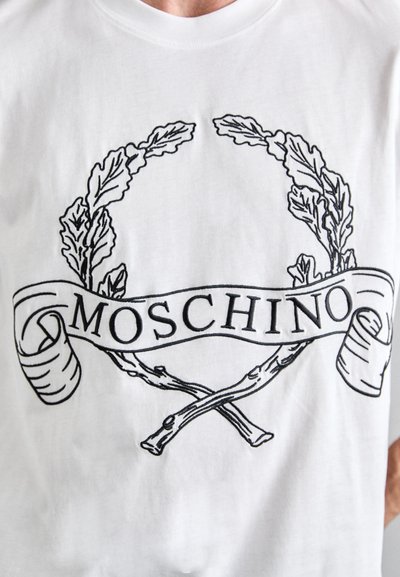 Bílý bavlněný tričko s černou grafikou vavřínových větví a stuhou se slovem "MOSCHINO" v tučném písmu.