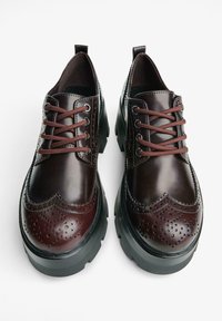 Un paio di scarpe brogue in pelle marrone scuro con suole nere spesse e lacci marroni, viste dall'alto su uno sfondo bianco.