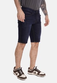 Shorts in denim blu scuro con taglio dritto, orli risvoltati, design a cinque tasche e dettagli di cucitura sottili. Abbinati a sneakers marroni e bianche.