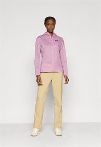 Ελαφρύ ροζ μπουφάν με κουκούλα, φερμουάρ μπροστά και λογότυπο στο στήθος, συνδυασμένο με μπεζ παντελόνι και κρεμ αθλητικά παπούτσια, σε απλό φόντο.