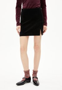 Schwarzer Wildleder-Minirock mit seitlichem Schlitz, kombiniert mit rot-schwarzen Strukturstrümpfen und schwarzen Schuhen mit einer dekorativen Schnalle.