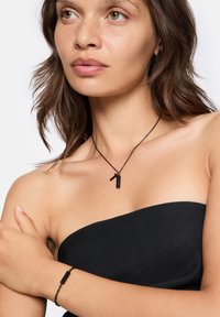 Sort halskæde med rektangulært vedhæng og matchende armbånd, begge med et slankt design og metallisk tekstur mod bar hud.