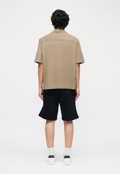 Άνδρας με σγουρά μαλλιά, γυρισμένος με την πλάτη, φοράει μπεζ πουκάμισο με κοντά μανίκια, μαύρο σορτς, λευκές κάλτσες και λευκά αθλητικά παπούτσια σε λευκό φόντο.