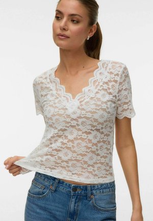 Mujer con top blanco de encaje con flores y mangas cortas, escote en V con bordes ondulados, combinado con jeans de mezclilla azul, sujetando el dobladillo del top.