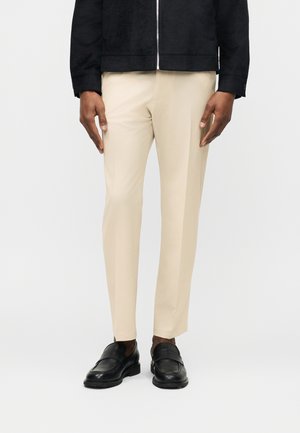 Man draagt beige op maat gemaakte broek en zwarte lederen loafers, met zichtbare mouwen van een zwarte jas, staand tegen een effen achtergrond.