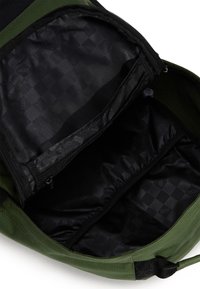 Interior de mochila de tela verde con forro negro, compartimentos con cremallera y acentos con patrón de diamantes. Presenta una textura suave y una forma estructurada.