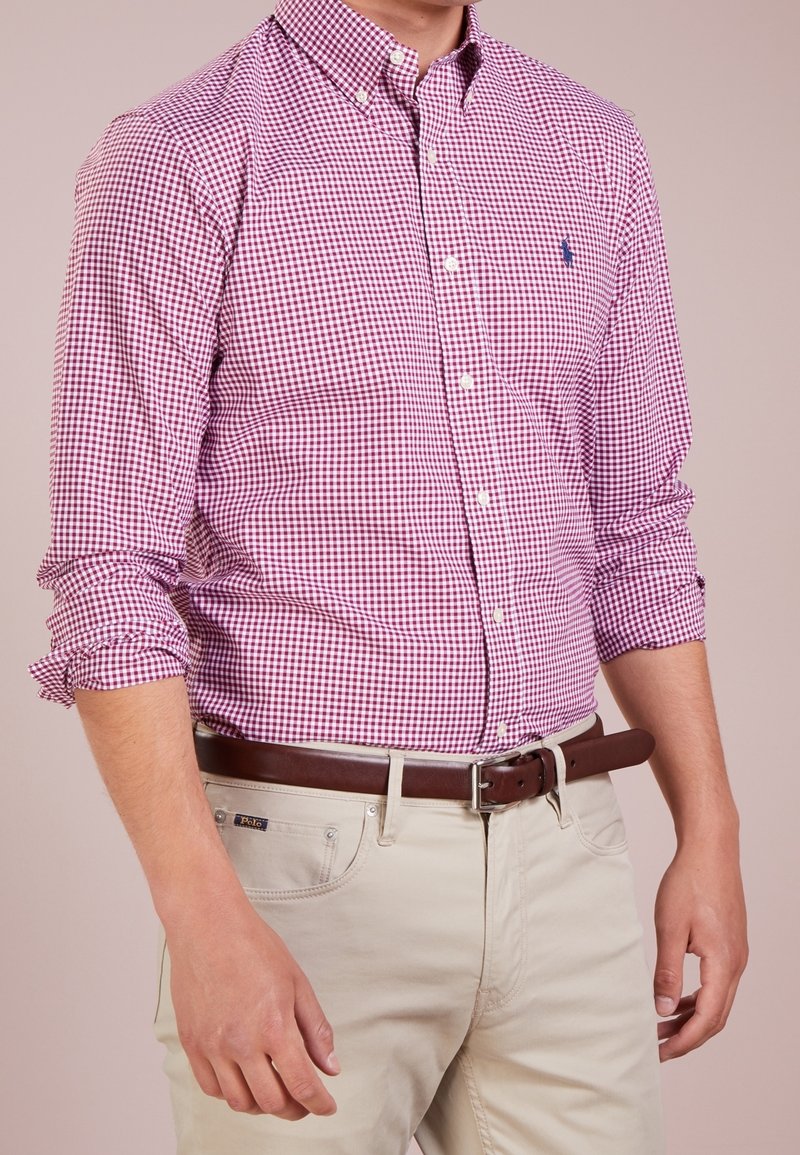 Czerwona i biała kraciasta koszula na guziki z długimi rękawami, podwiniętymi mankietami i małym logo. Noszona z jasnymi spodniami i brązowym paskiem.