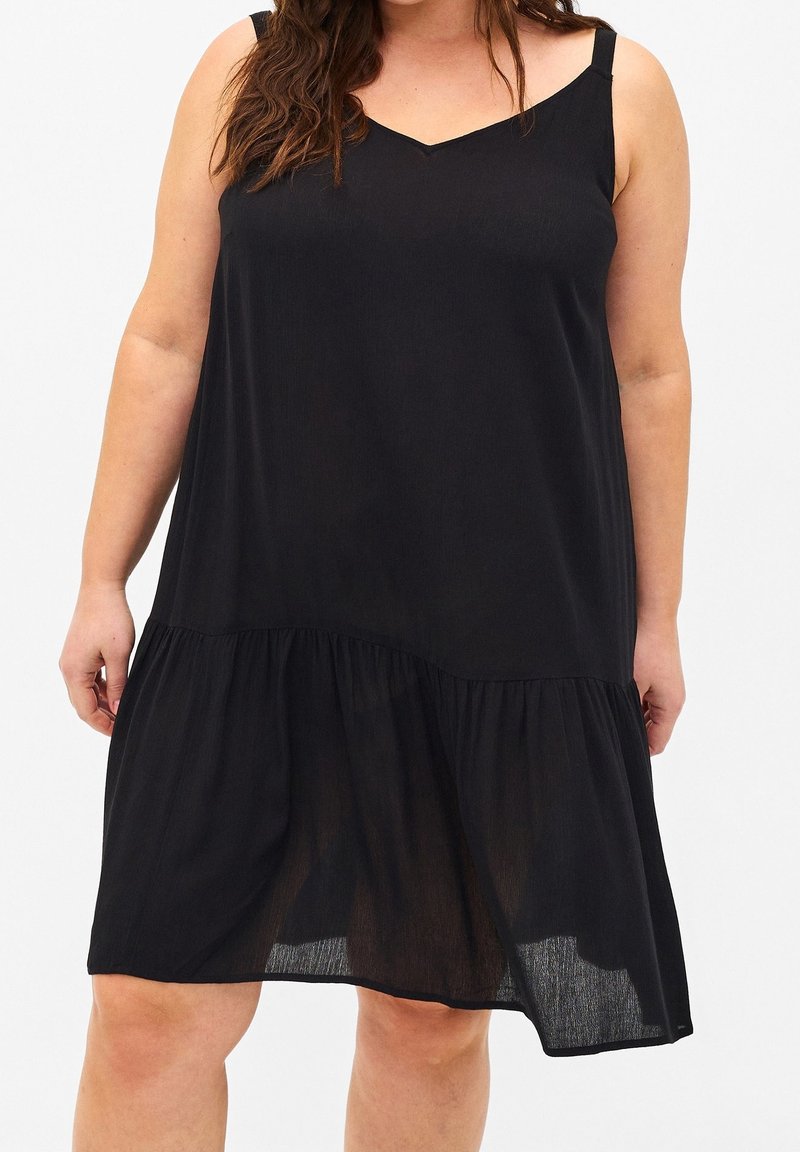 Žena v bezramenných černých šatech ke kolenům s volánovým lemem, stojící na jednobarevném bílém pozadí.