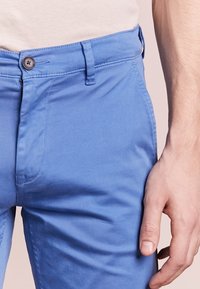 Ljusblå bomullshängselbyxor med dragkedja och knappstängning, som har bälteslänkar och ett slätt, något texturerat tyg.