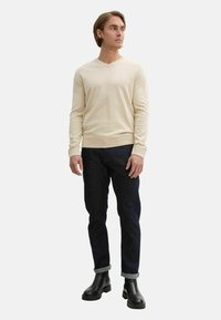 Beiger V-Ausschnitt-Pullover, blaue Denim-Jeans, schwarze Leder-Stiefeletten, die eine taillierte Silhouette mit gerippten Bündchen und Saum zeigen.