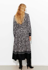 Robe noire et blanche à motifs paisley, avec manches longues, taille cintrée et bordure inférieure noire, assortie à des bottes noires montantes.