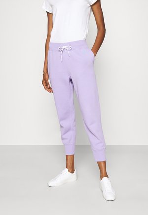 Osoba nosząca lawendowe spodnie dresowe ze białym sznurkiem, białą koszulkę i białe trampki, stojąca z jedną ręką w kieszeni.