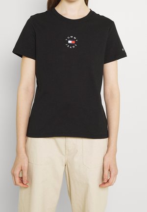 Person iført en sort Tommy Jeans t-shirt med logo på brystet og cremefarvede bukser står mod en ensfarvet hvid baggrund.