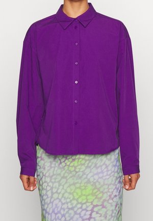 Fioletowa koszula na długi rękaw, zapinana na guziki, z szpiczastym kołnierzykiem, noszona na zielonej i lawendowej spódnicy z abstrakcyjnym wzorem.