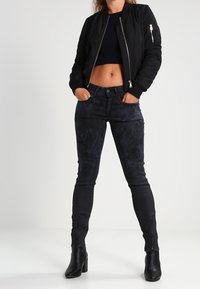 Chaqueta bomber negra con cremallera en el frente, top ajustado azul marino y vaqueros skinny negros con un acabado texturizado. Botas de tacón negras completan el conjunto.