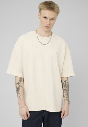 Jeune homme portant un t-shirt crème ample et un jean foncé, montrant des tatouages de rose et de papillon sur les deux avant-bras, avec des cheveux blonds courts.