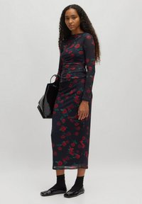 Zwarte lange mouwen jurk met rode bloemenprint, aansluitende silhouette, gerimpelde details en midi-lengte. Gepaard met een zwarte tas en platte schoenen.