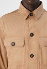 Veste en daim beige avec un col pointu, deux poches poitrine et des boutons noirs. Texture douce avec un design simple et structuré.
