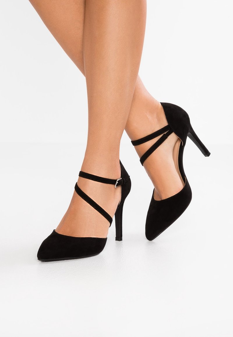 Jambes portant des escarpins noirs en daim à bout pointu avec des doubles sangles chevillant croisées sur les pieds, posés sur un sol blanc.
