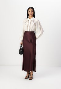 Weiße Bluse mit Cut-Out-Details und puffärmel, kombiniert mit einem burgunderfarbenen Satinrock. Eine schwarze Handtasche und filigrane Sandalen runden das Outfit ab.