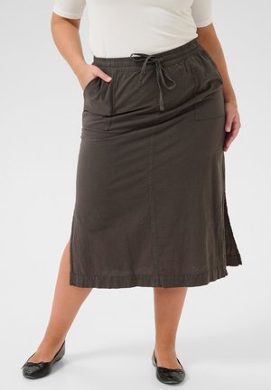 Kvinde iført en mørk nederdel med lommer, der går til knæene, med snor i taljen, kombineret med sorte flade sko og en hvid top med korte ærmer.