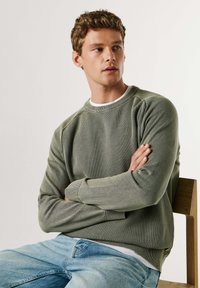 Roheline tekstuuriga pusakangas ümar kaelusega, soonikkoes varrukaservade ja kitseneva lõikega, kombineeritud helesiniste teksadega. Modelli seljas, kes istub puidust toolil.