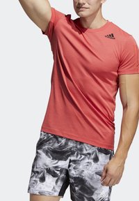 Rød atletisk t-shirt med tekstureret design og korte ærmer, parret med sort-hvide mønstrede shorts. Har brandlogo på brystet.