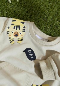 Beige huppari, jossa on tiikerigrafiikka ja sana "RAWR" mustana. Varustettu ribbineuloksella loppuilla ja pehmeällä tekstuurilla. Ainulaatuinen muotoiluhyödyke olkapäällä.