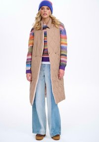 Beżowa pikowana kamizelka bez rękawów zapinana na zatrzaski, noszona na wielokolorowym swetrze w paski. Jasnoniebieskie szerokie spodnie i brązowe buty na platformie.