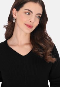 Fekete bordázott pulóver V-nyakkivágással és finom kötött mintákkal. Hosszú, hullámos barna haj kiegészíti a megjelenést, amely fémes fülbevalókat is tartalmaz.