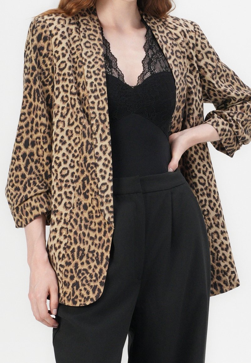 Blazer à imprimé léopard en tissu doux, avec une coupe décontractée, des manches trois-quarts et un col châle, superposé sur un bodysuit noir.