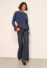 Tumedast sinise pikakä sleeves blouse, millel on volditud vöökoht, koos laia säärega tumedate teksade ja mustade terava ninaga kontsadega, hoides burgundi värvi vutlarit.