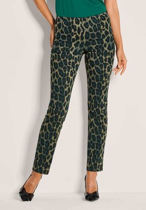 Frau in schmal geschnittenen Leopardenprint-Hosen und schwarzen spitzen High Heels, die vor einer schlichten hellen Wand steht.