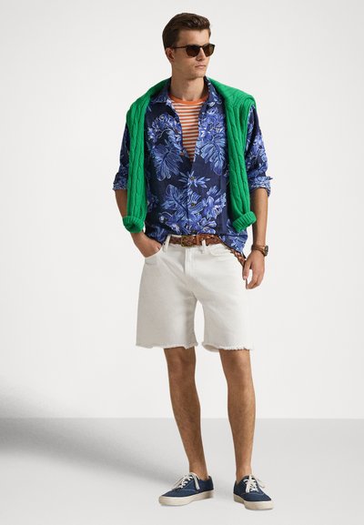 Άνδρας με γυαλιά ηλίου, μπλε πουκάμισο με λουλούδια πάνω από πορτοκαλί ριγέ μπλουζάκι, πράσινο πουλόβερ περασμένο στους ώμους, λευκές βερμούδες και μπλε αθλητικά παπούτσια.