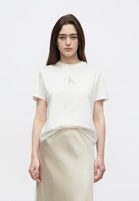 Balta kokvilnas T-krekls ar īsām piedurknēm un apaļu kakla izliekumu, uz krūtīm izvietots gaiši rozā 'CK' logotips, kopā ar bēša krāsas svārkiem.